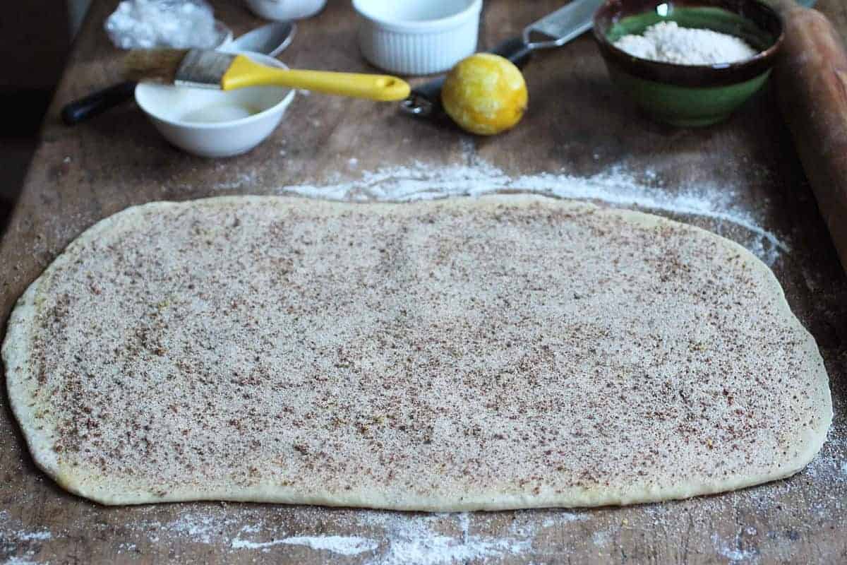 Rolled rectangle of dough with cinnamon sugar on a wooden table; bowls and lemon in the background.