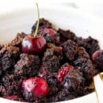Close up of chocolate dump cake with cherries in a white bowl, a spoon, white surface