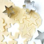 On a white surface rolled cookie dough with cut outs and metal shapes, red text on top