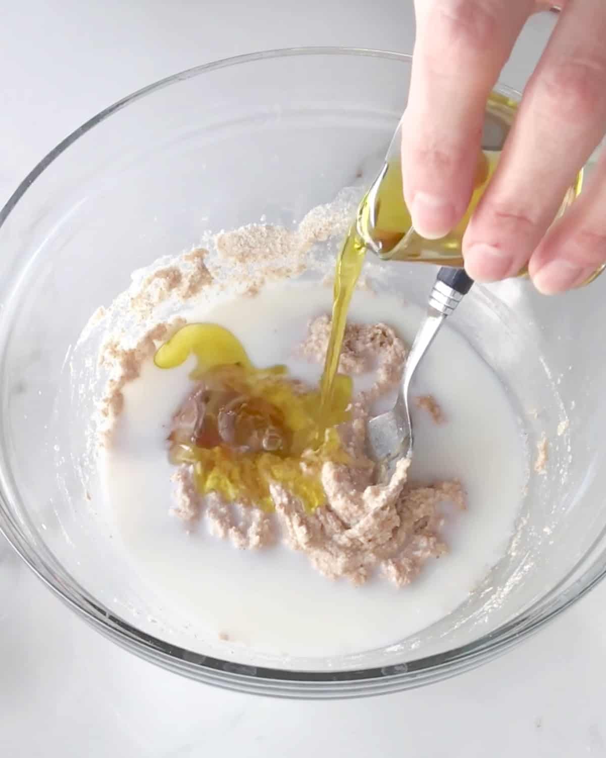 Milk and oil added to whole wheat dough in a glass bowl. White marble surface.