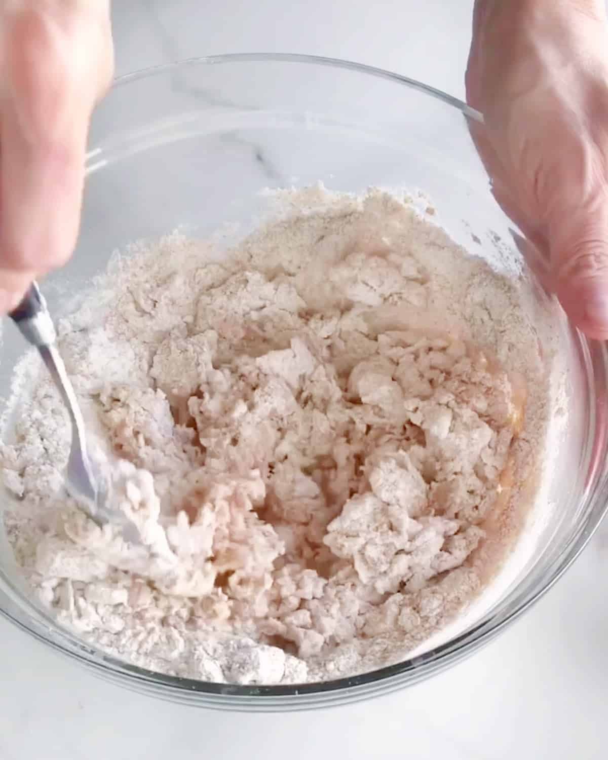 Whole wheat flour added to bread dough in a glass bowl. Hands mixing it. White marble surface.