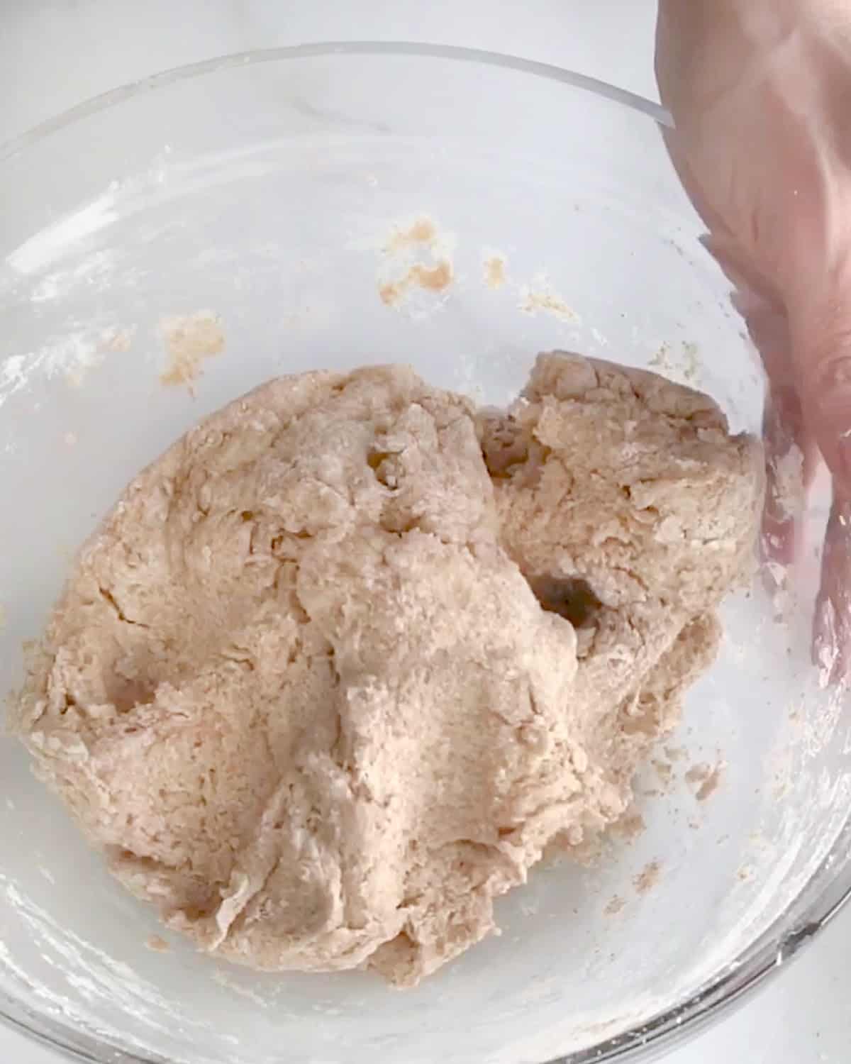 Stiff whole wheat bread dough in a glass bowl on a white surface.