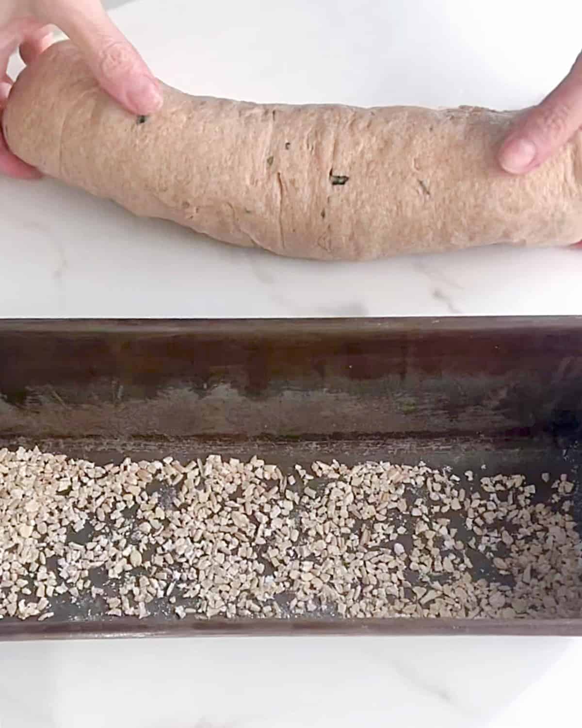 Loaf of bread and a loaf pan with oats in the bottom on a white marble surface.