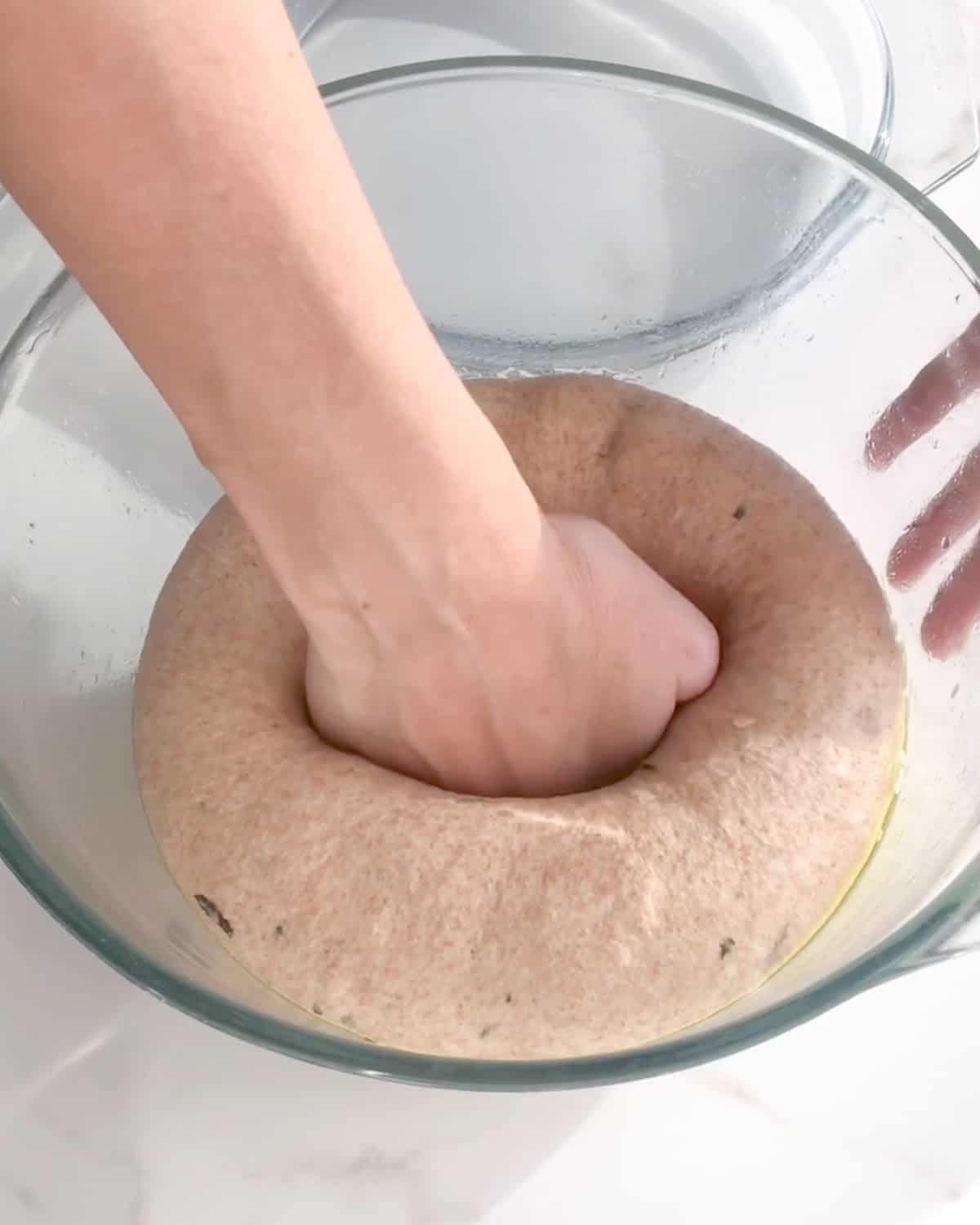 Hand deflating a whole wheat bread dough in a glass bowl on a white surface.