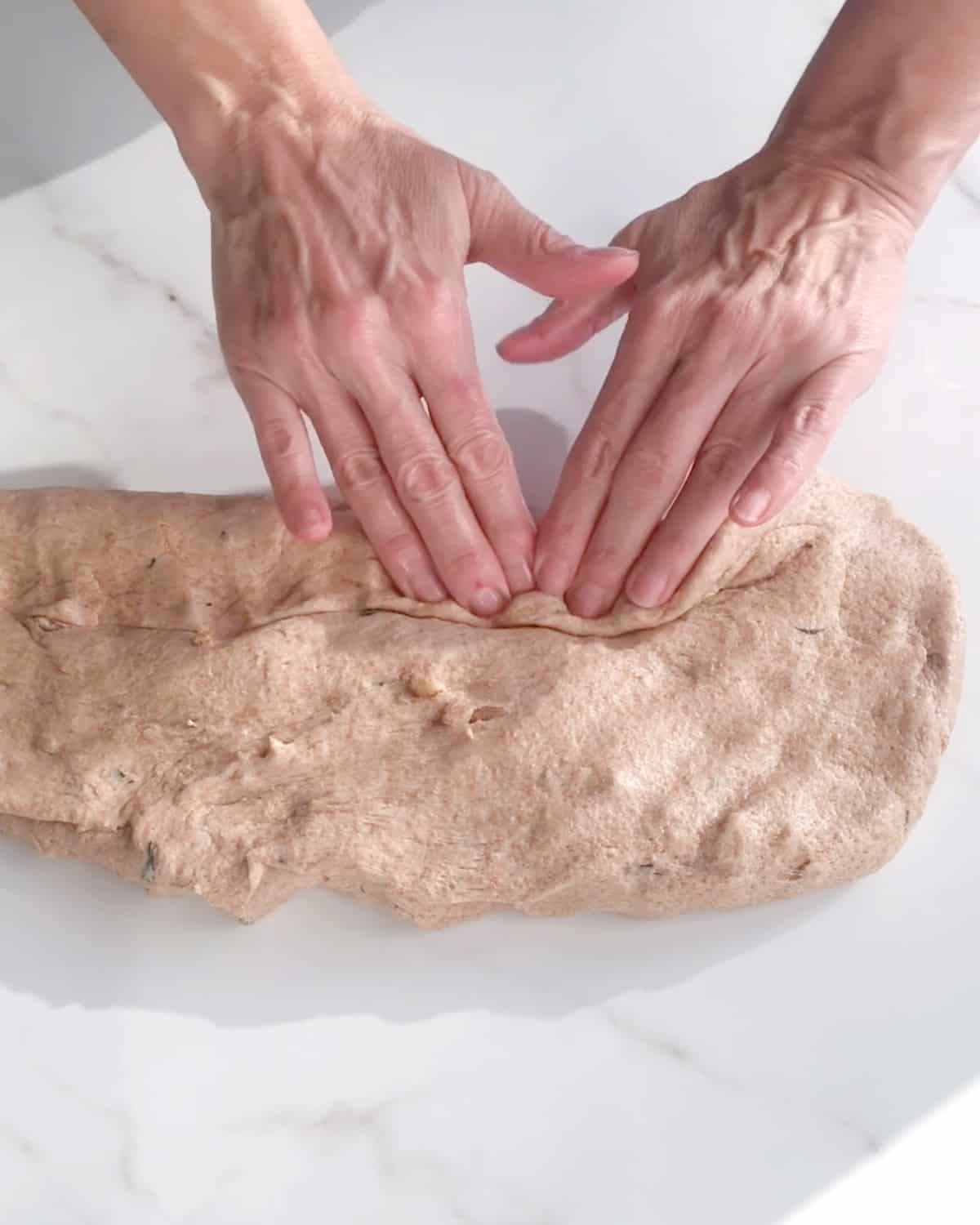 Hands forming a whole wheat bread loaf on a white marble surface.