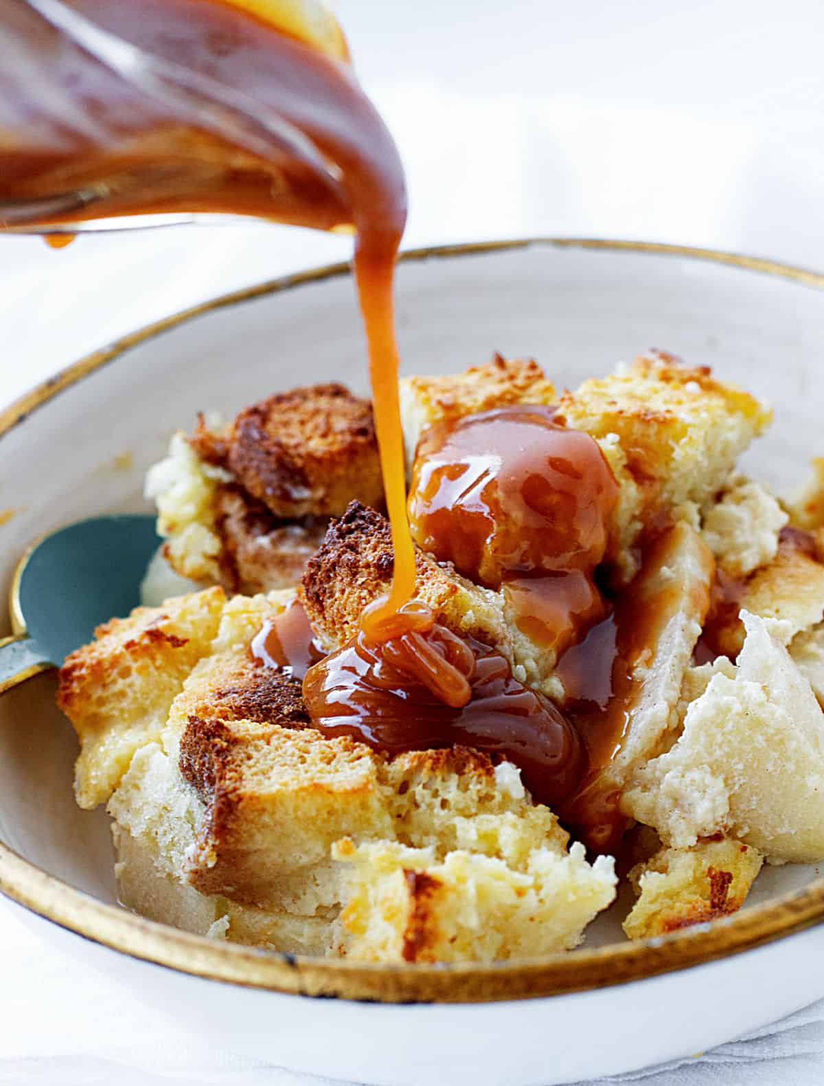 Pouring dulce de leche on serving of apple bread pudding, white bowl and background.