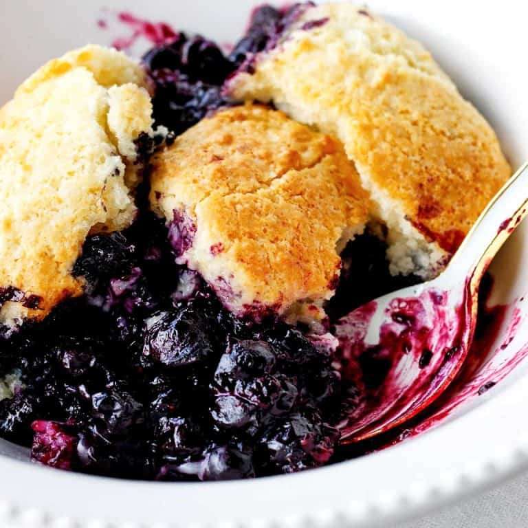 A white bowl and light blue spoon with serving of blueberry cobbler. Close up image.