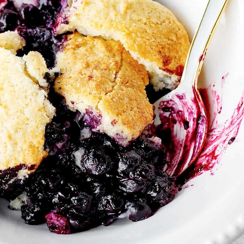 Serving of blueberry cobbler in a white bowl with a light blue spoon. Close up image.