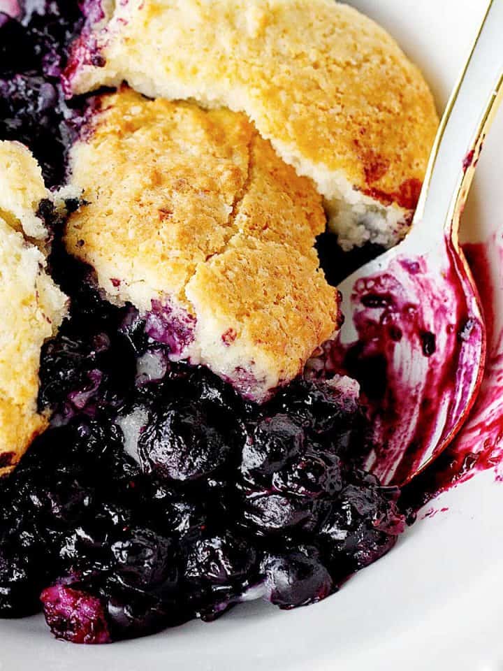 Serving of blueberry cobbler in a white bowl with a light blue spoon. Close up image.