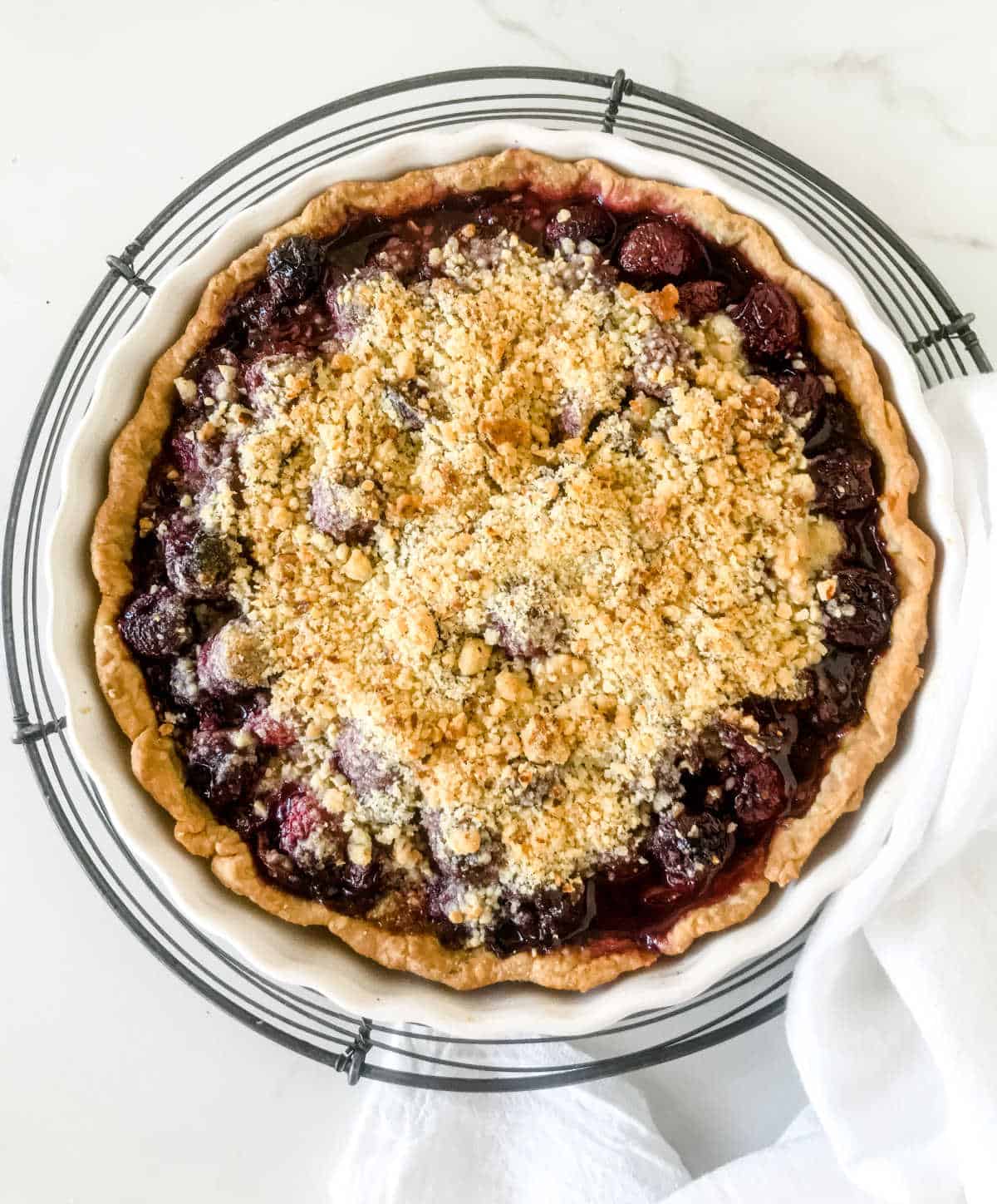 Top view of whole cherry crumb pie on wire rack, white towel and surface
