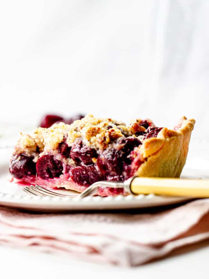 White background and plate with cherry pie slice, a fork, light pink napkin underneath