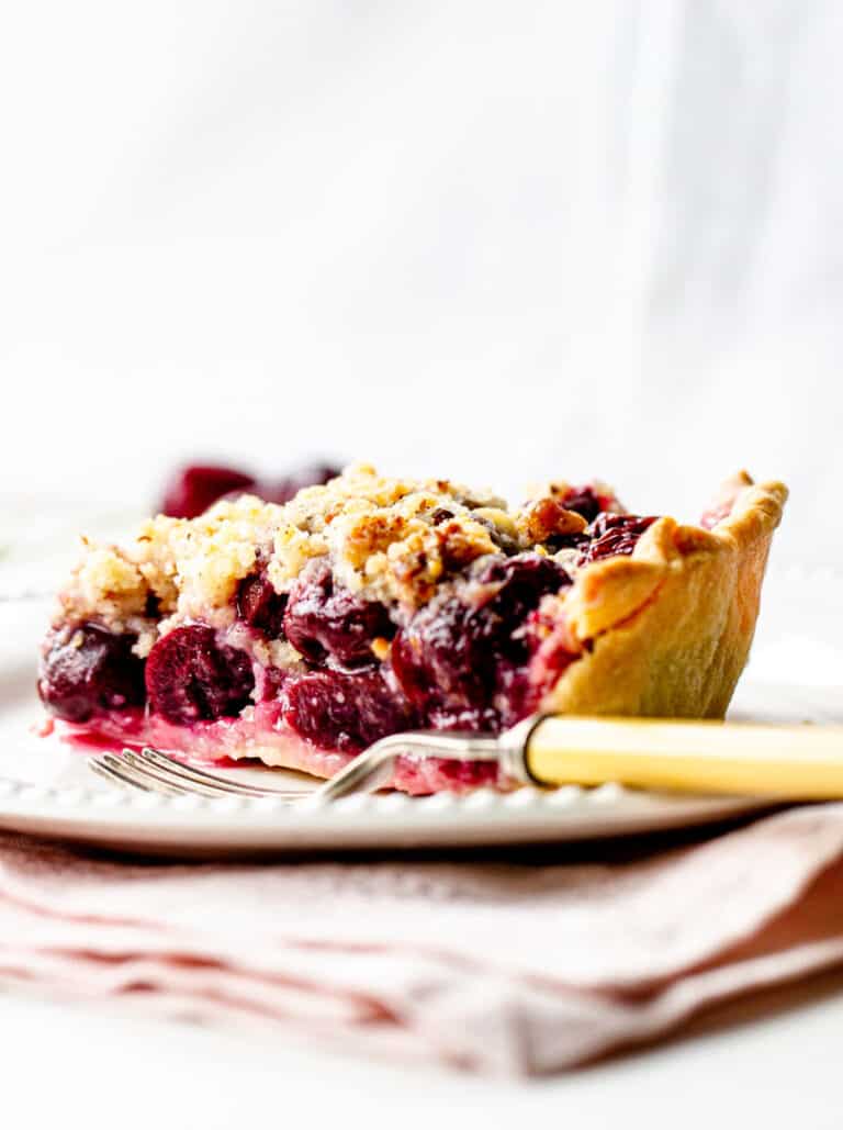 White background and plate with cherry pie slice, a fork, light pink napkin underneath