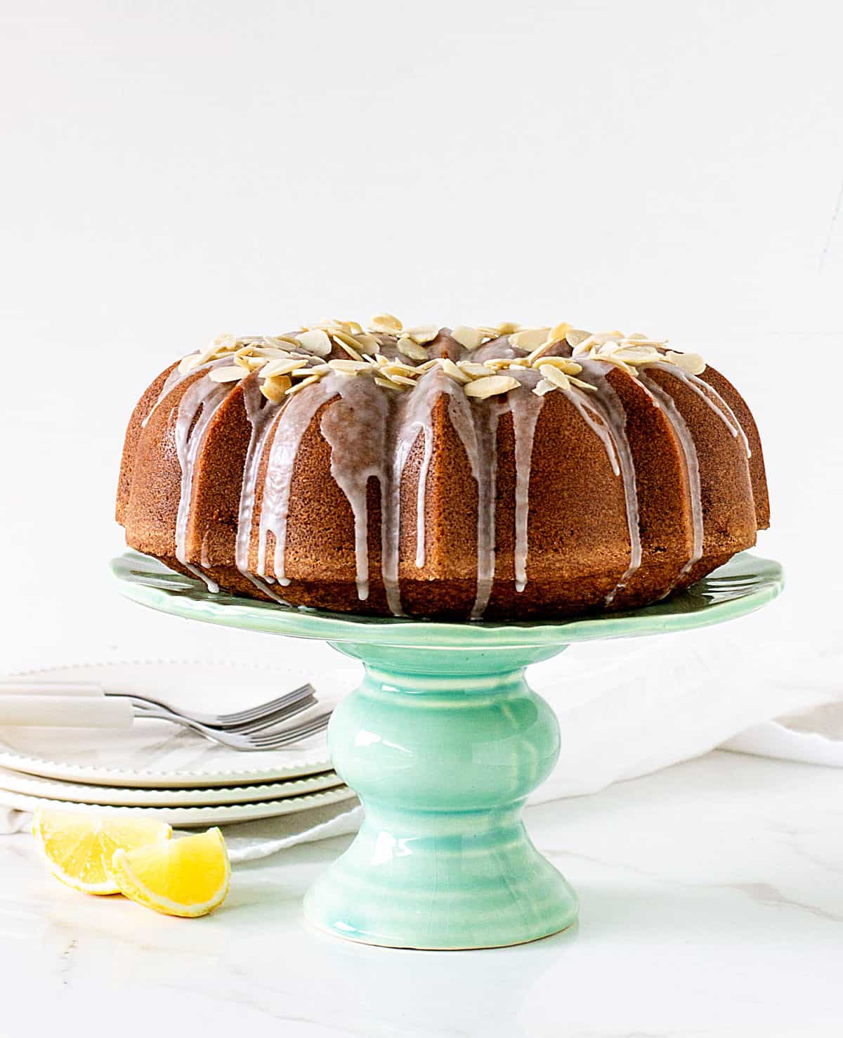 Front view of whole almond topped bundt cake on green cake stand, white plates and background, lemon wedges