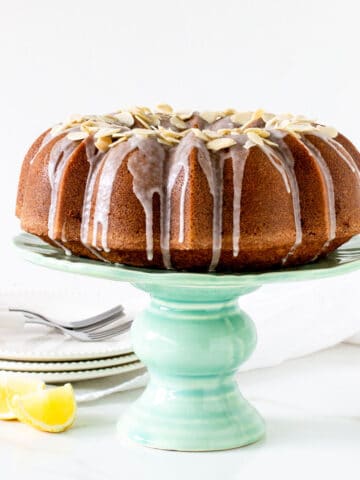 A whole bundt cake on a green cake stand, white background, lemon wedges, plate stack