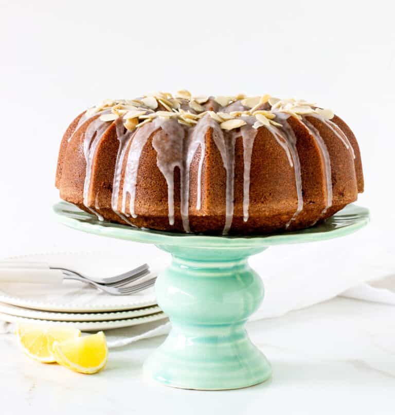 A whole bundt cake on a green cake stand, white background, lemon wedges, plate stack