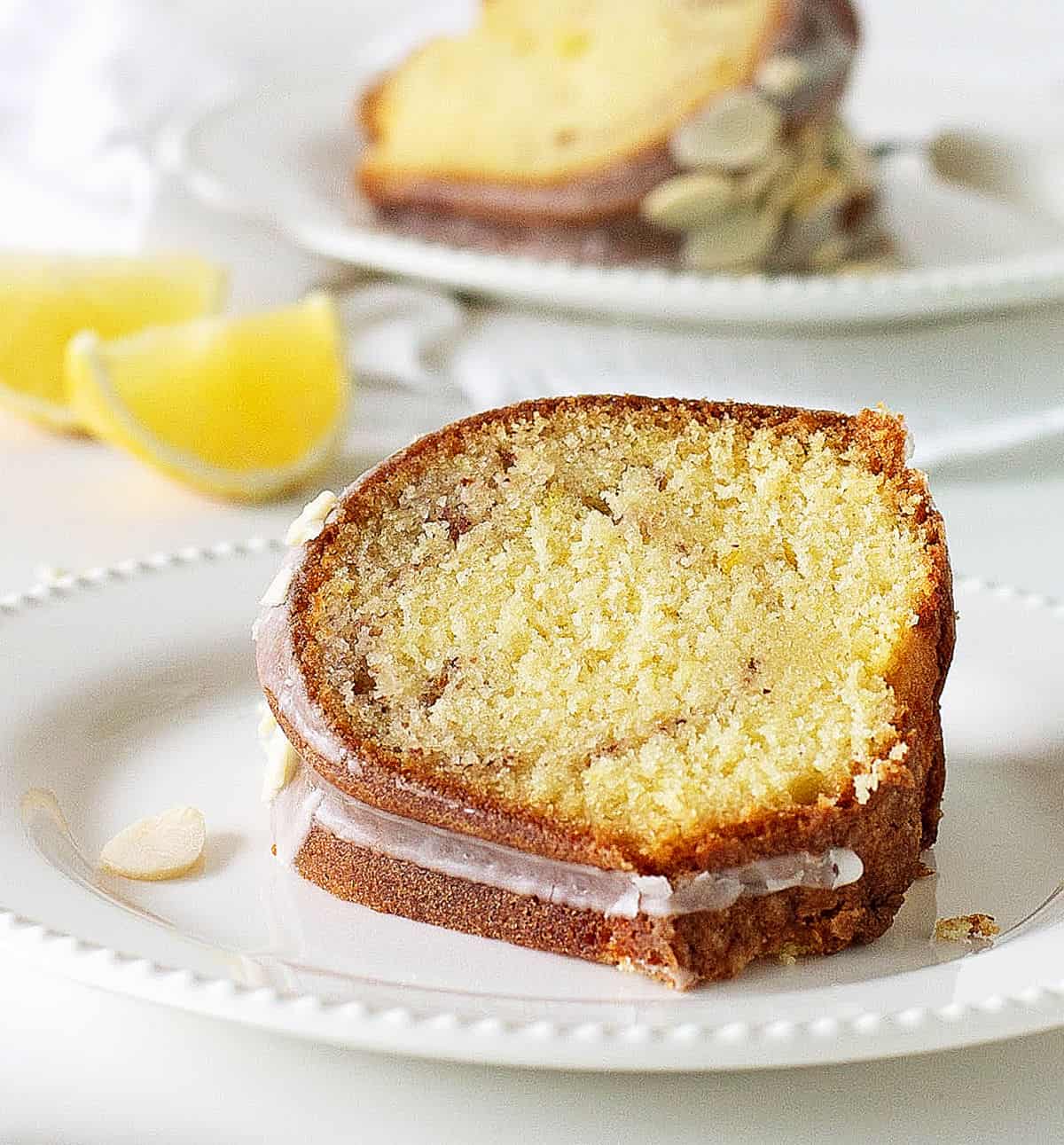 Slice of lemon bundt cake on white plate, white background with more cake and lemon wedges