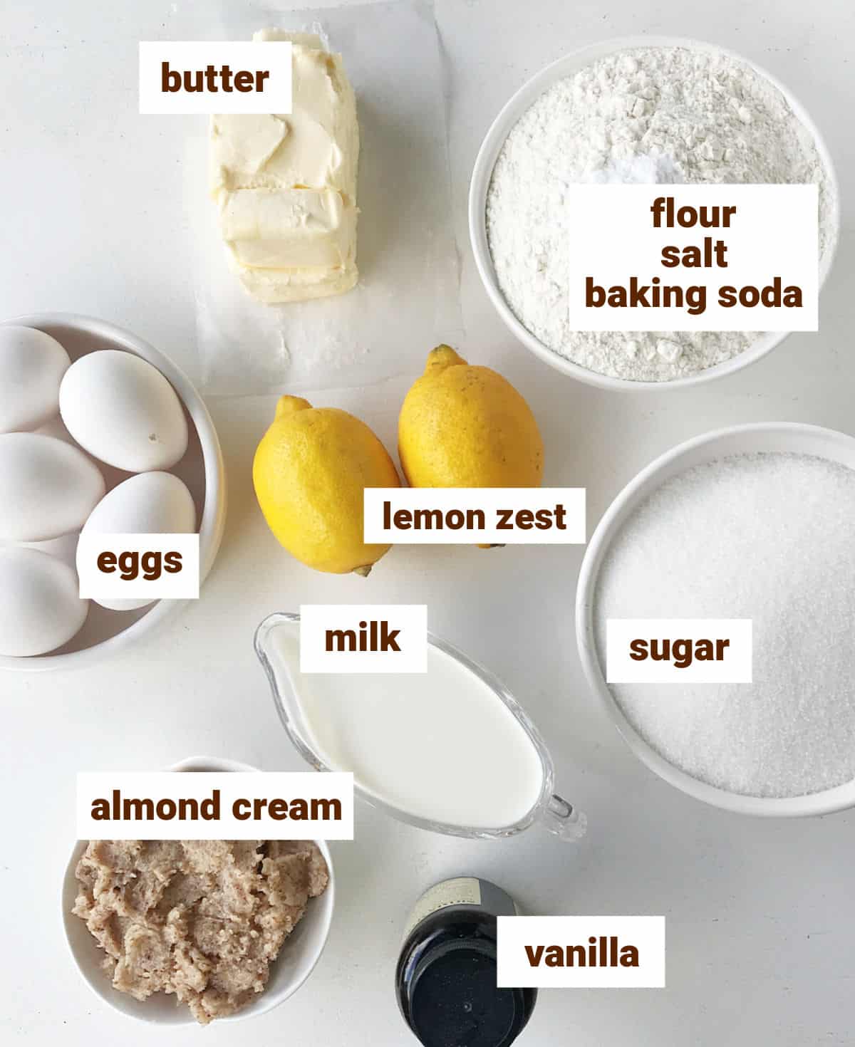 Bowls with ingredients for lemon almond cake, including milk, vanilla, eggs, butter on a white surface