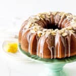 Partial view of almond topped glazed bundt cake on greenish cake stand, white background and surface, lemon wedges