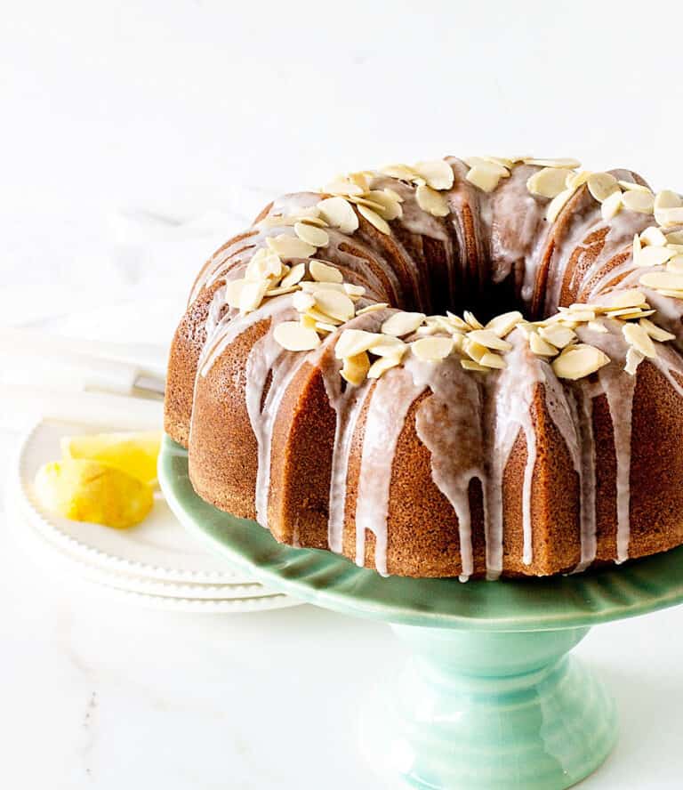 Partial view of almond topped glazed bundt cake on greenish cake stand, white background and surface, lemon wedges