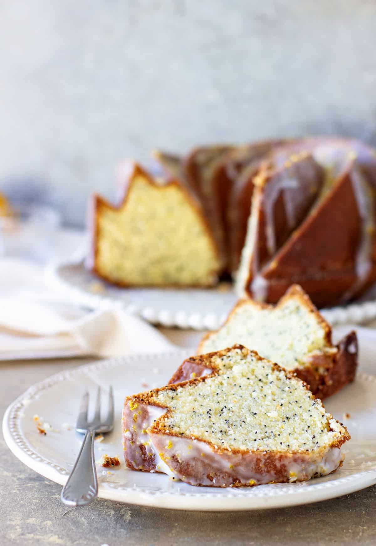 Slices of lemon poppy seed bundt cake on white plate with fork, cake in background.