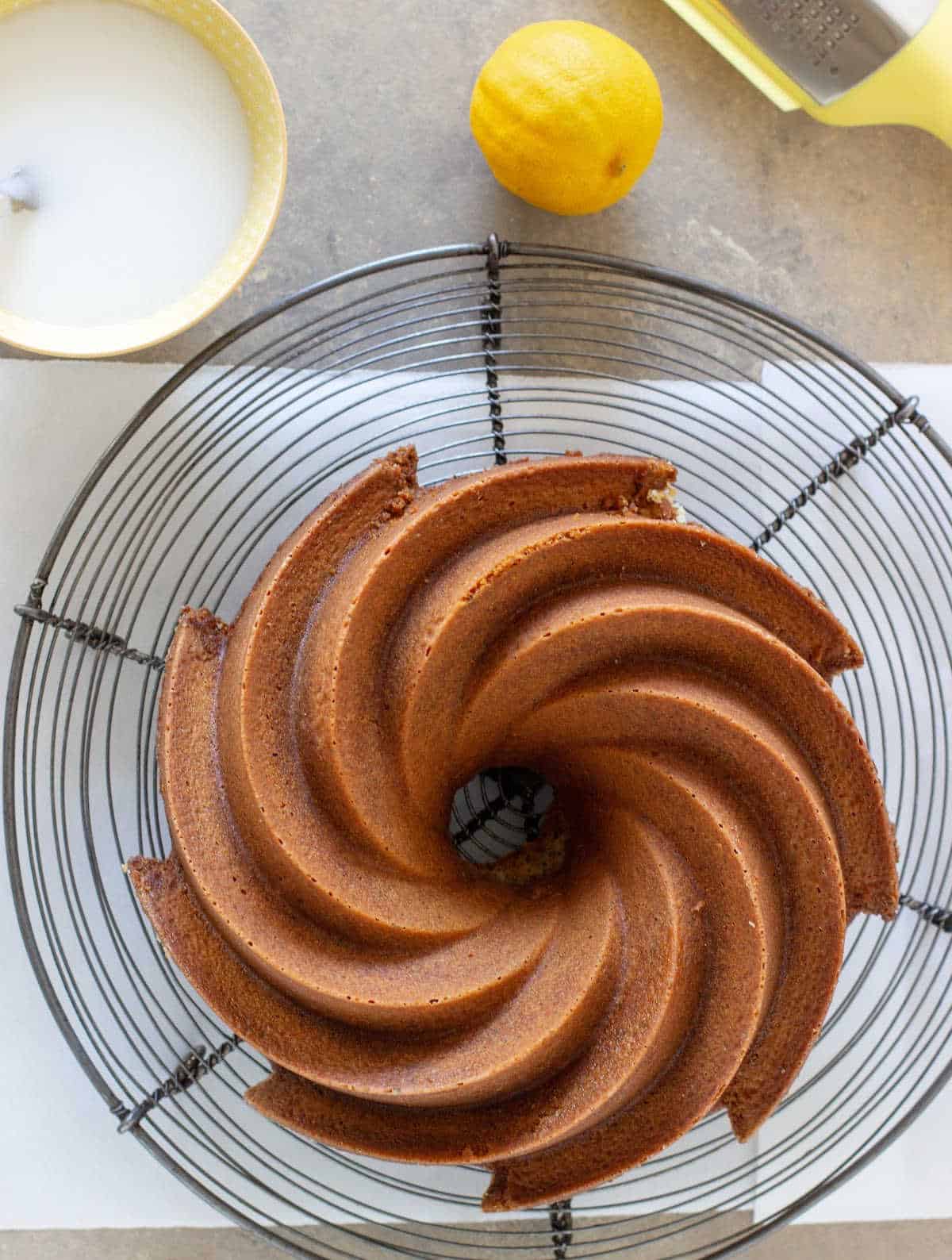Wire rack and parchment paper with bundt cake, bowl with glaze, a lemon and grater. Grayish surface.