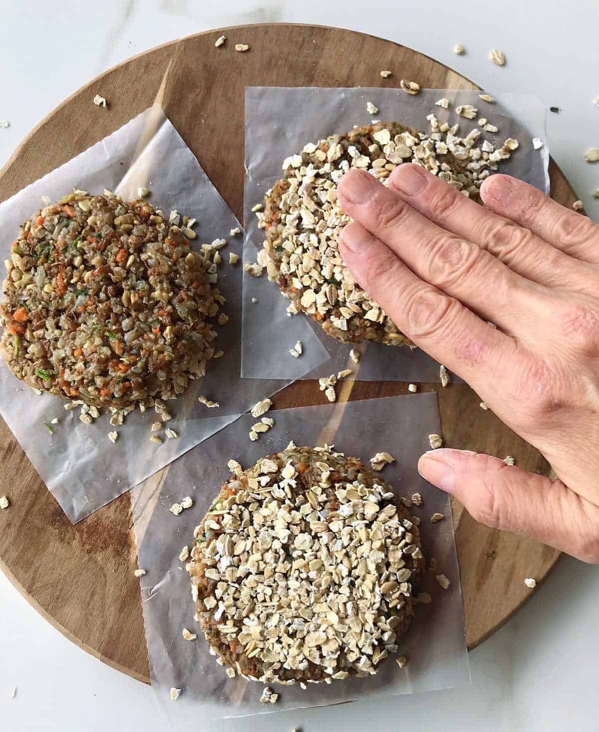 Hand pressing oats onto veggie burgers on a round wooden board