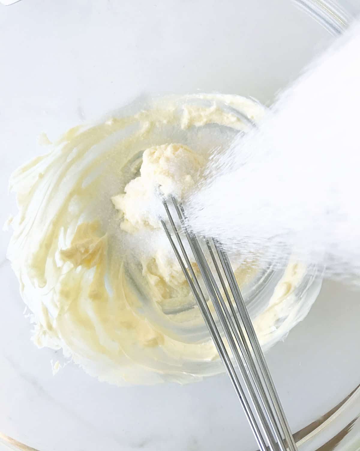 Adding sugar to butter in a glass bowl with a hand whisk inside