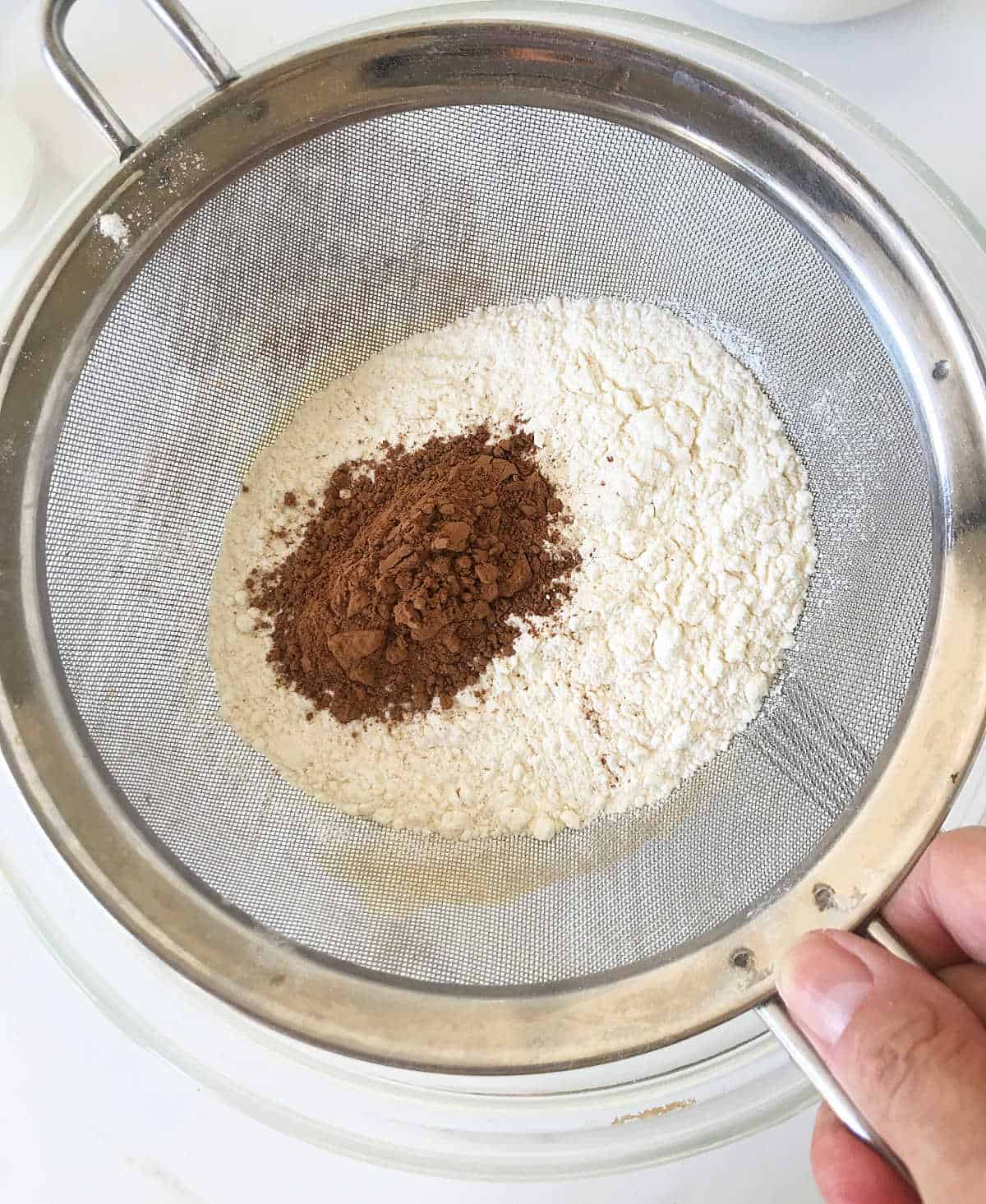 Sifter with flour and cocoa powder on top of glass bowl
