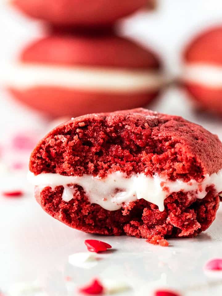 Single bitten red velvet whoopie pie on white marble surface. More pies in the background.