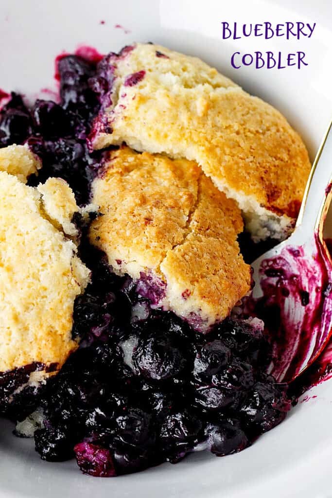 Serving of blueberry cobbler on white bowl, a spoon; purple text overlay