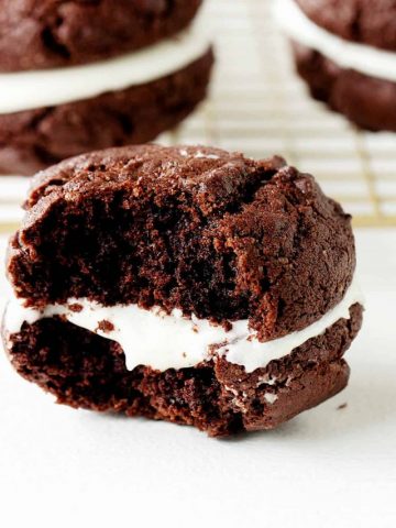 Close up of half a chocolate whoopie with cream filling, white surface, gold wire rack, more pies