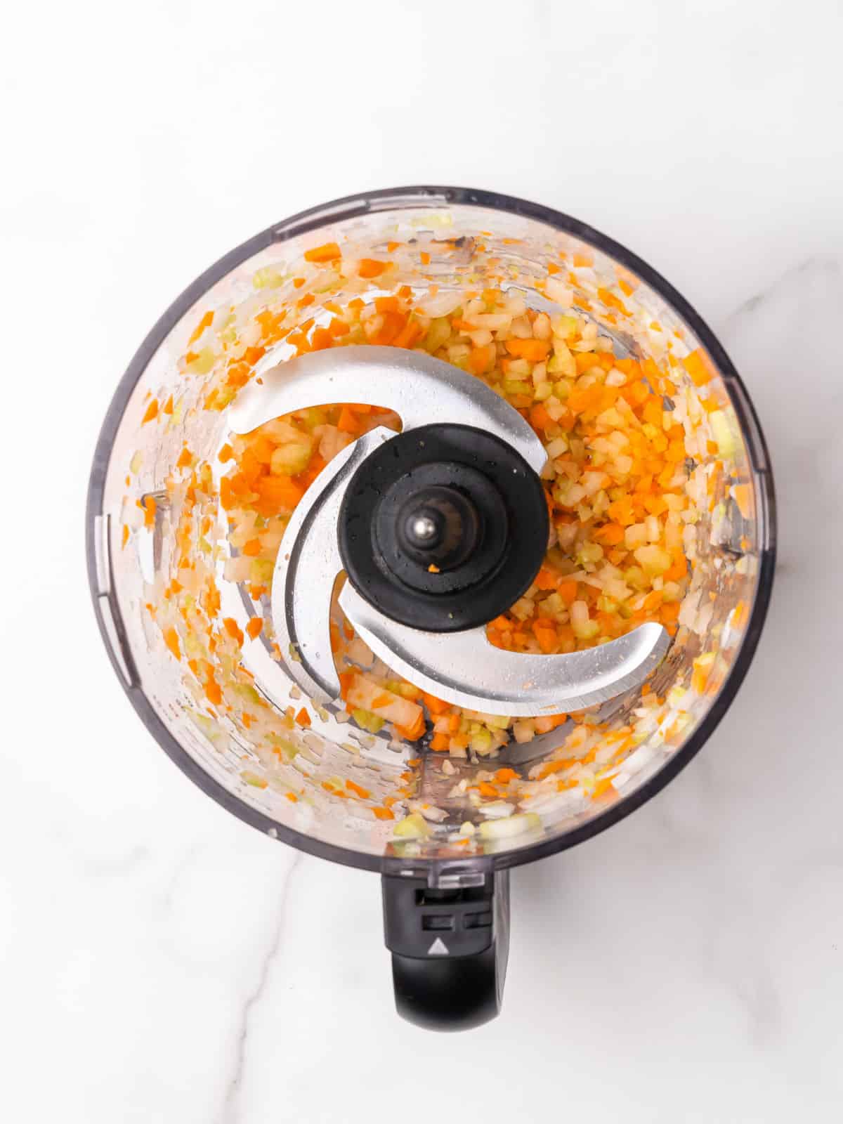 Top view of food processor bowl with chopped carrot, celery, and onion. White marble surface.