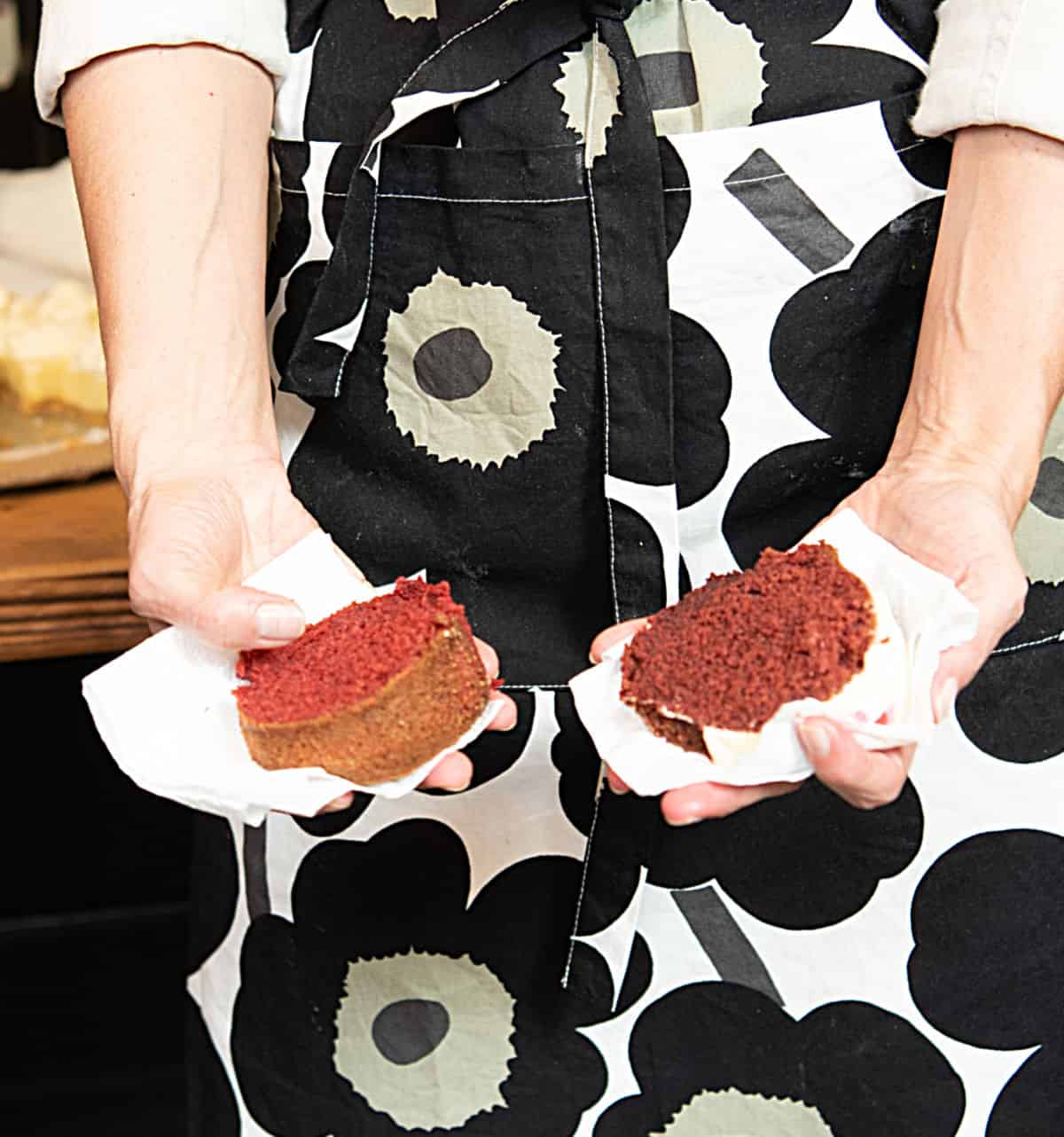 Holding slices of red velvet cake, one in each hand, dark flowered apron