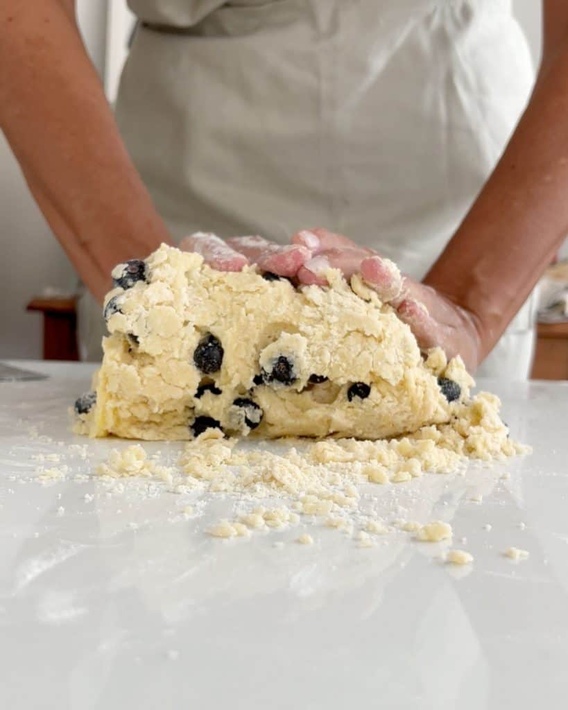Front view of person integrating a blueberry scone dough on white marble surface.