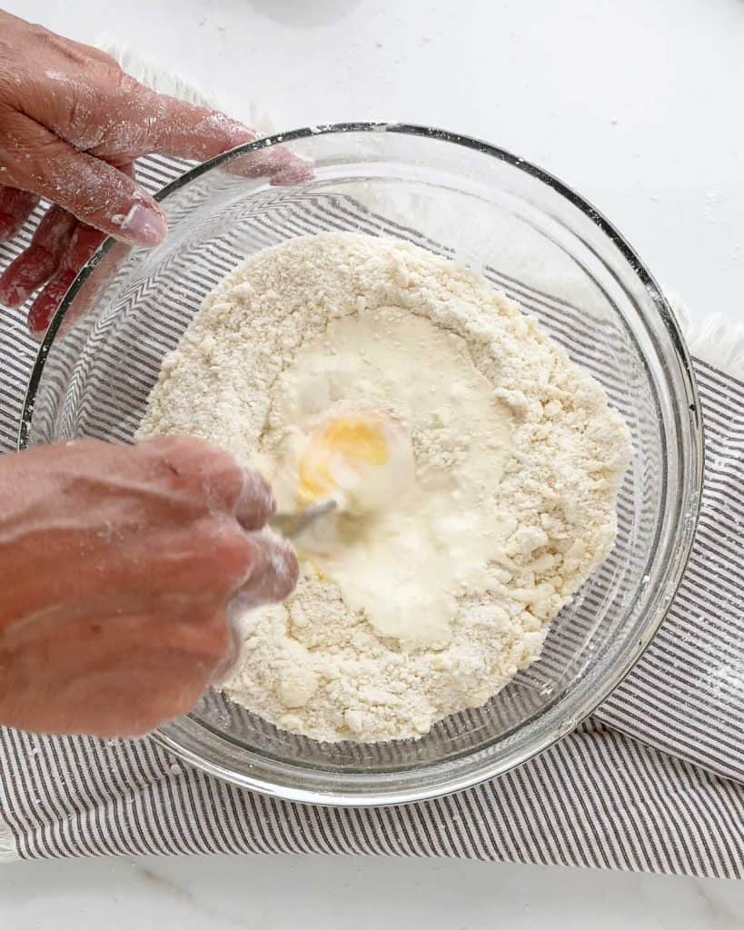 Mixing egg and milk with dry ingredients in a glass bowl on a blue striped cloth on a white surface.