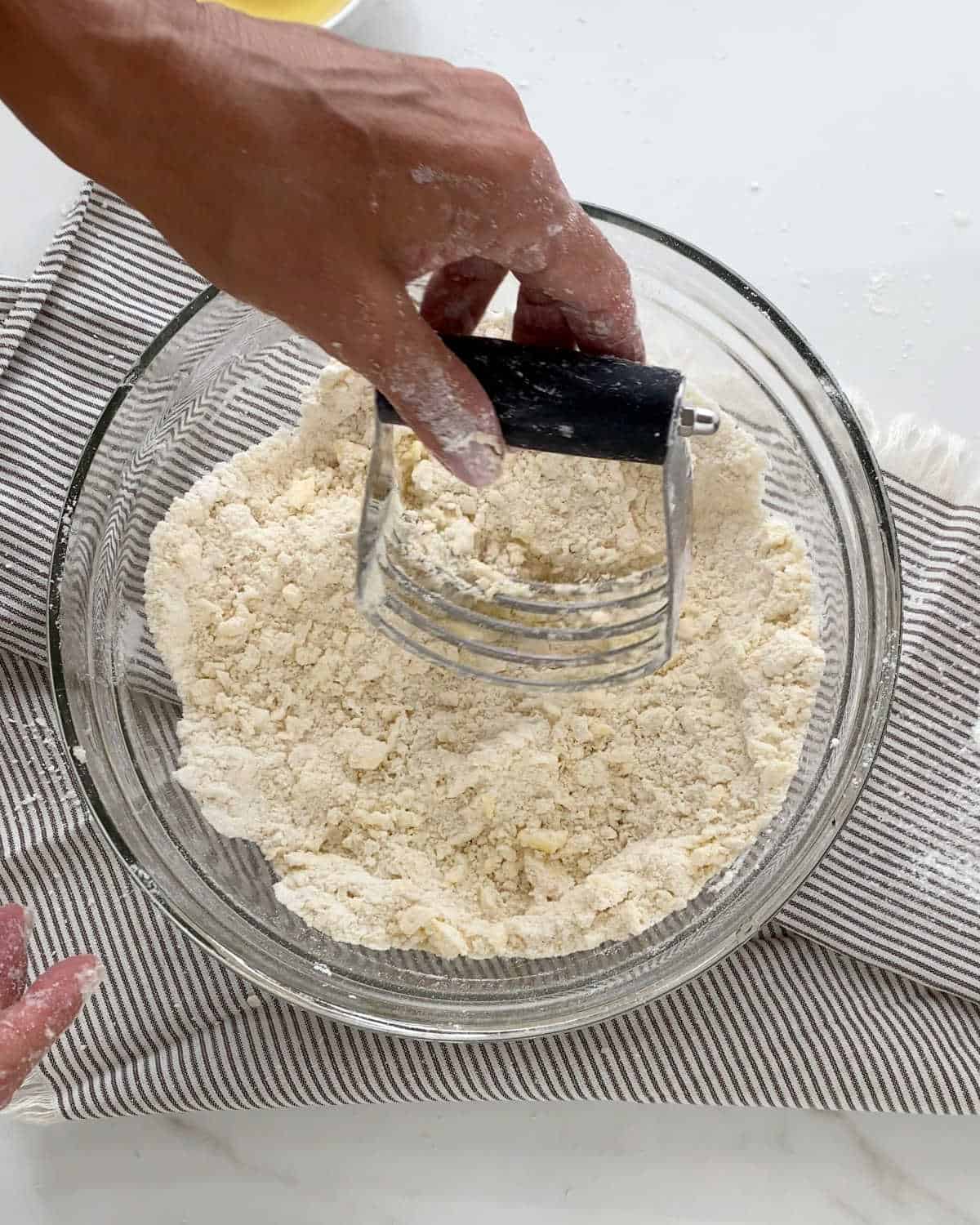Cutting scone dough in a glass bowl with a pastry cutter. White marble and striped cloth beneath the bowl. 