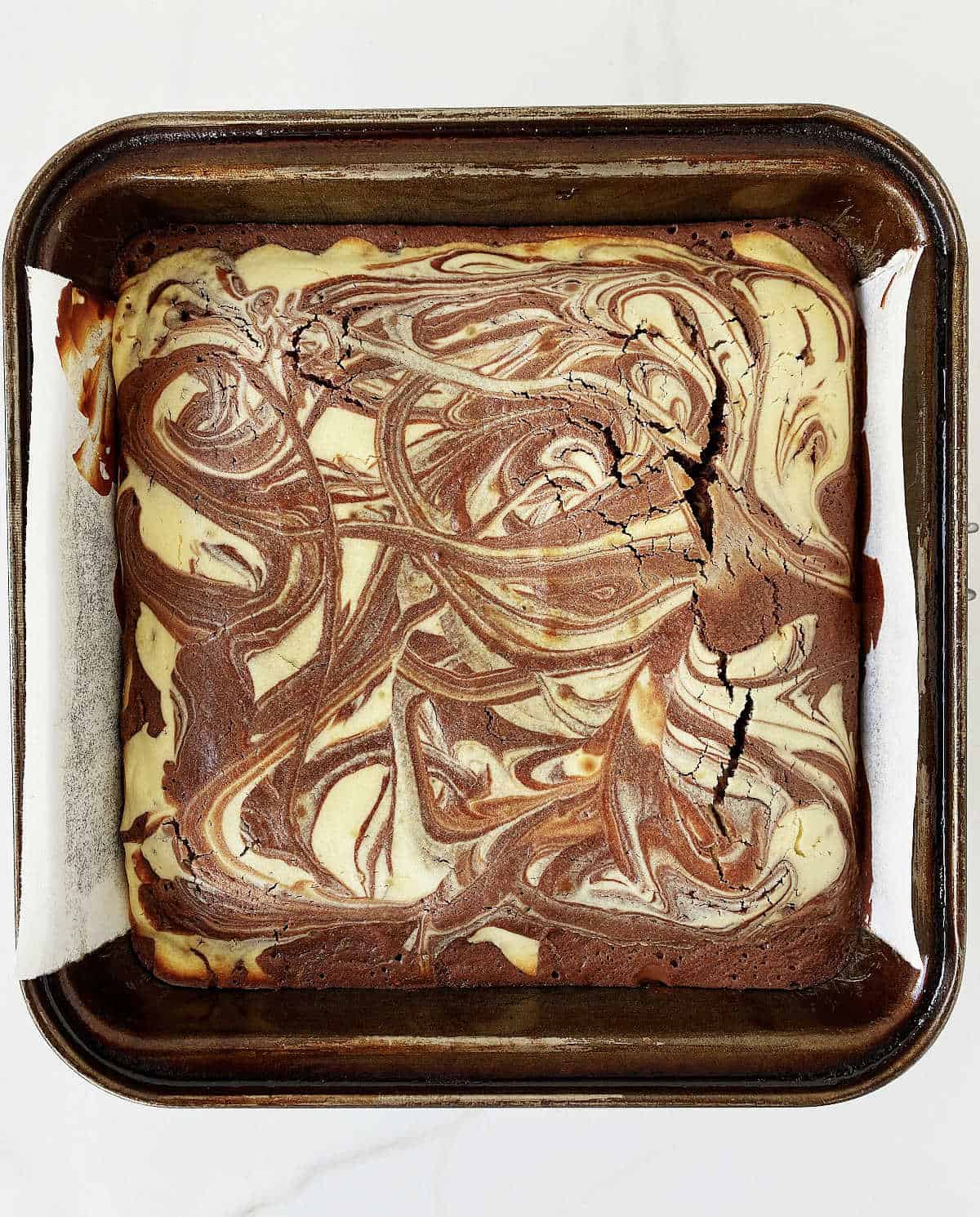 Baked cheesecake brownies in a metal square pan on white marble.