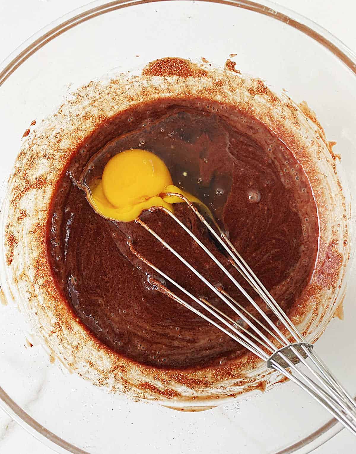 Adding egg to brownie batter in a glass bowl with a whisk.