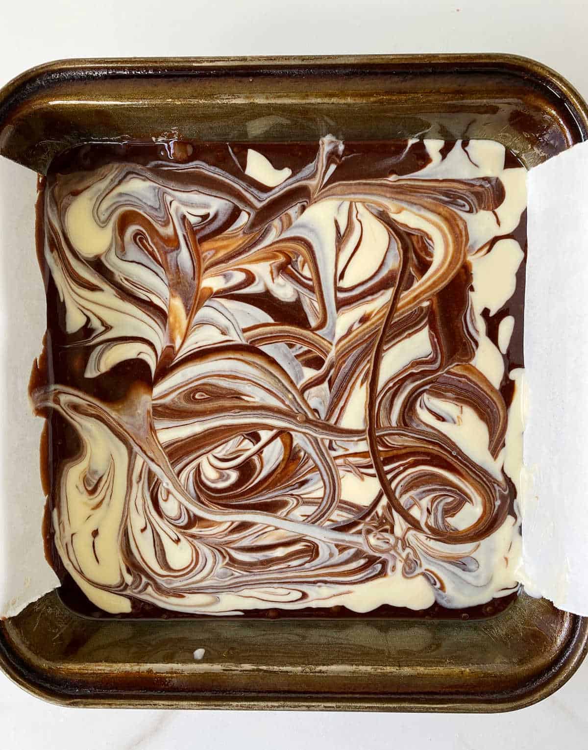 Brownie batter with cheesecake swirl on a metal square pan on a white surface. 
