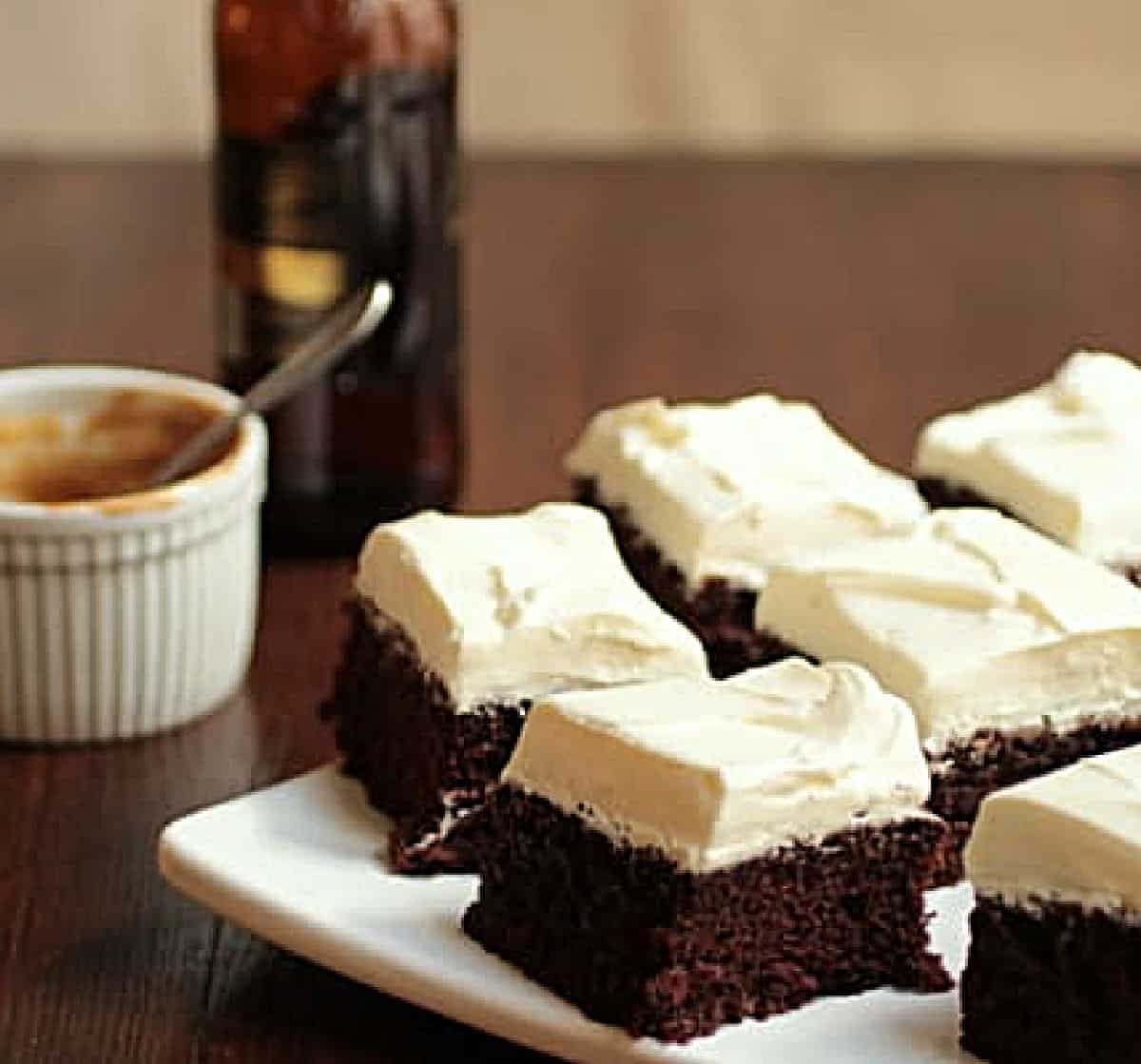 Cream topped chocolate cake squares on white plate, wooden table, stout bottle and white ramekin around