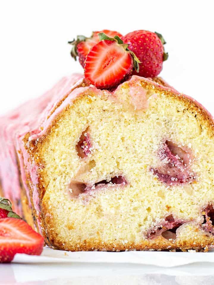 A cut loaf of strawberry pound cake, with a pink glaze and fresh strawberries. White background.