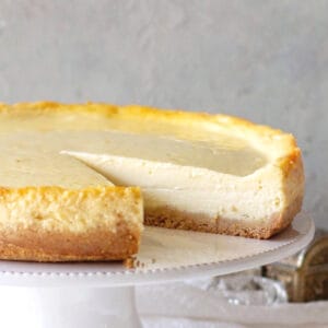 Plain cheesecake missing a slice on a white cake stand, grey background