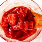 Wooden spoon showing stawberry sauce, glass bowl beneath and white surface