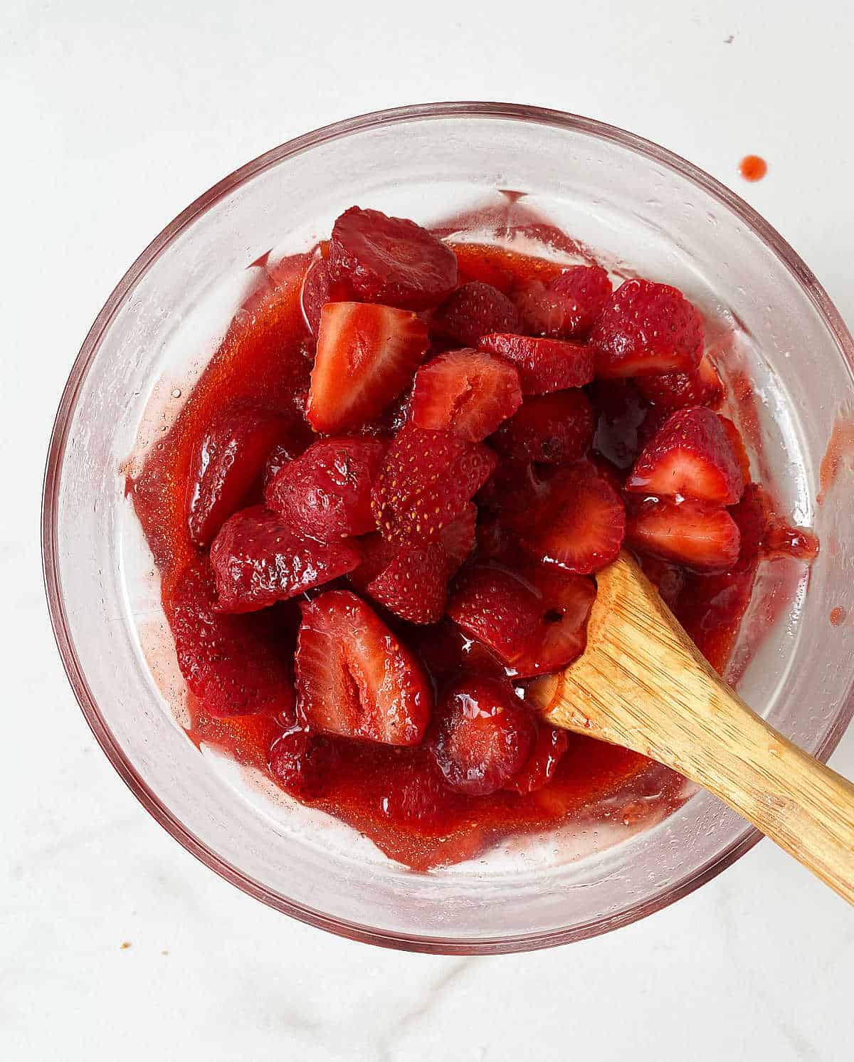 Glass bowl with strawberry sauce and fresh chopped strawberries, a wooden spoon inside, white surface