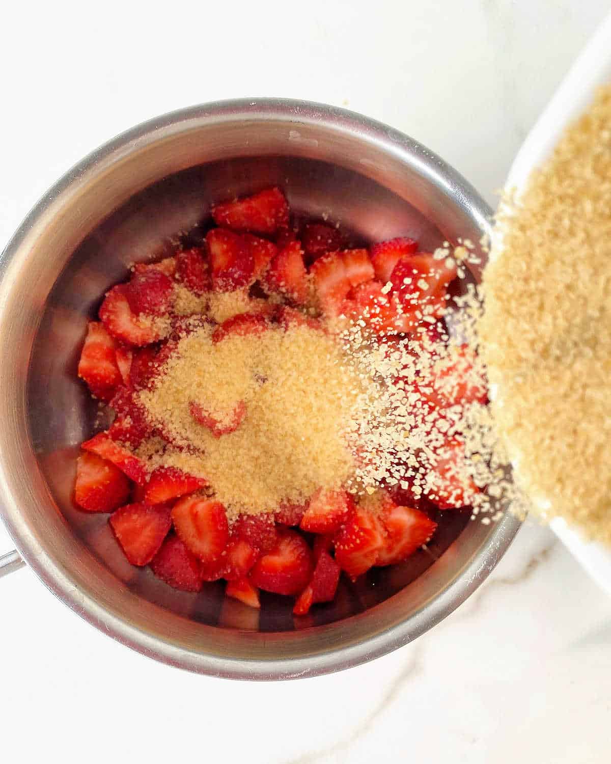 Adding brown sugar to metal saucepan with strawberry pieces. White surface.