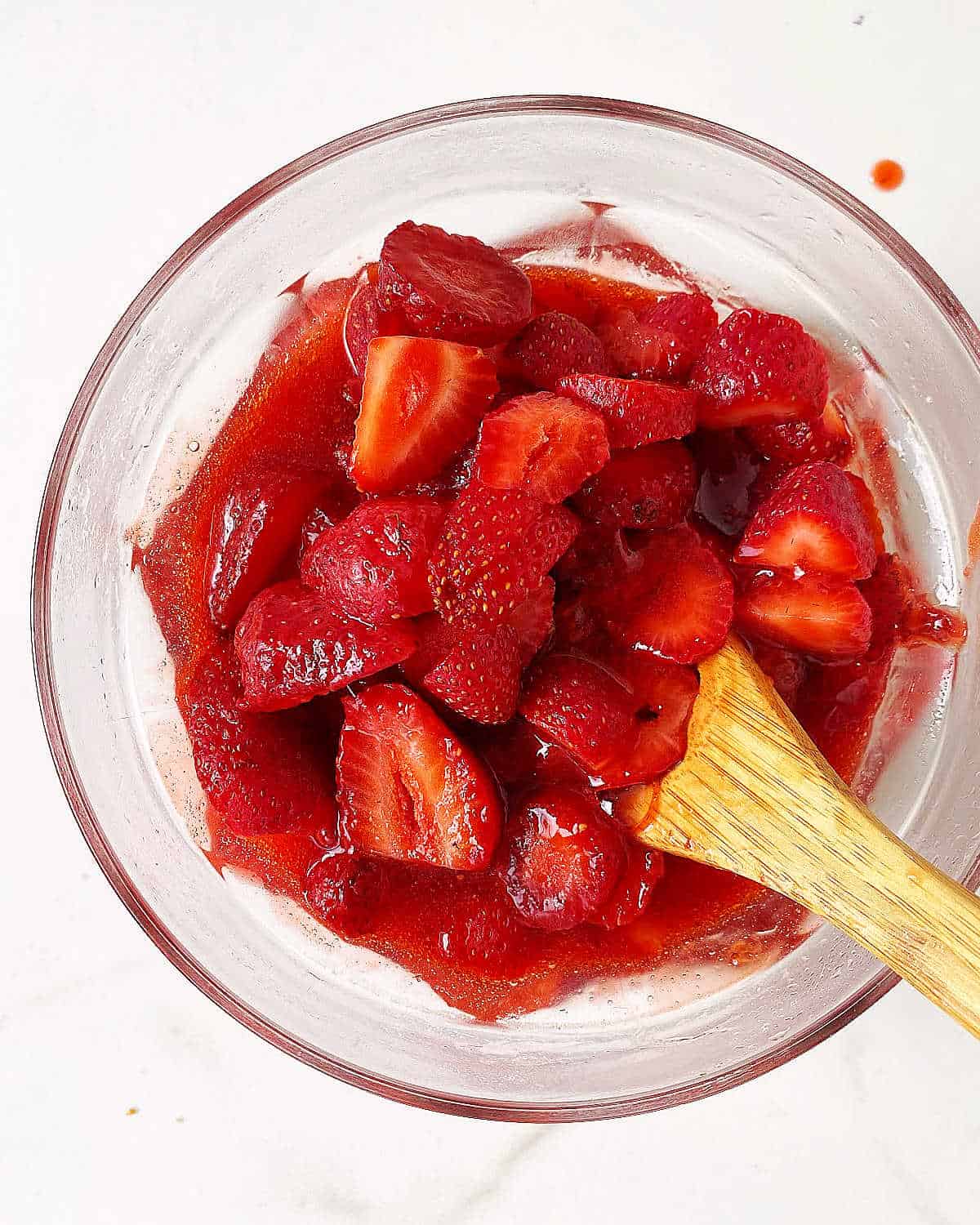 Glass bowl with strawberry sauce and fresh chopped strawberries, a wooden spoon inside, white surface.