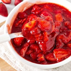 A white napkin with a white bowl of strawberry sauce being spooned. Fresh scattered strawberries.