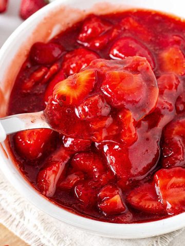 A white napkin with a white bowl of strawberry sauce being spooned. Fresh scattered strawberries.