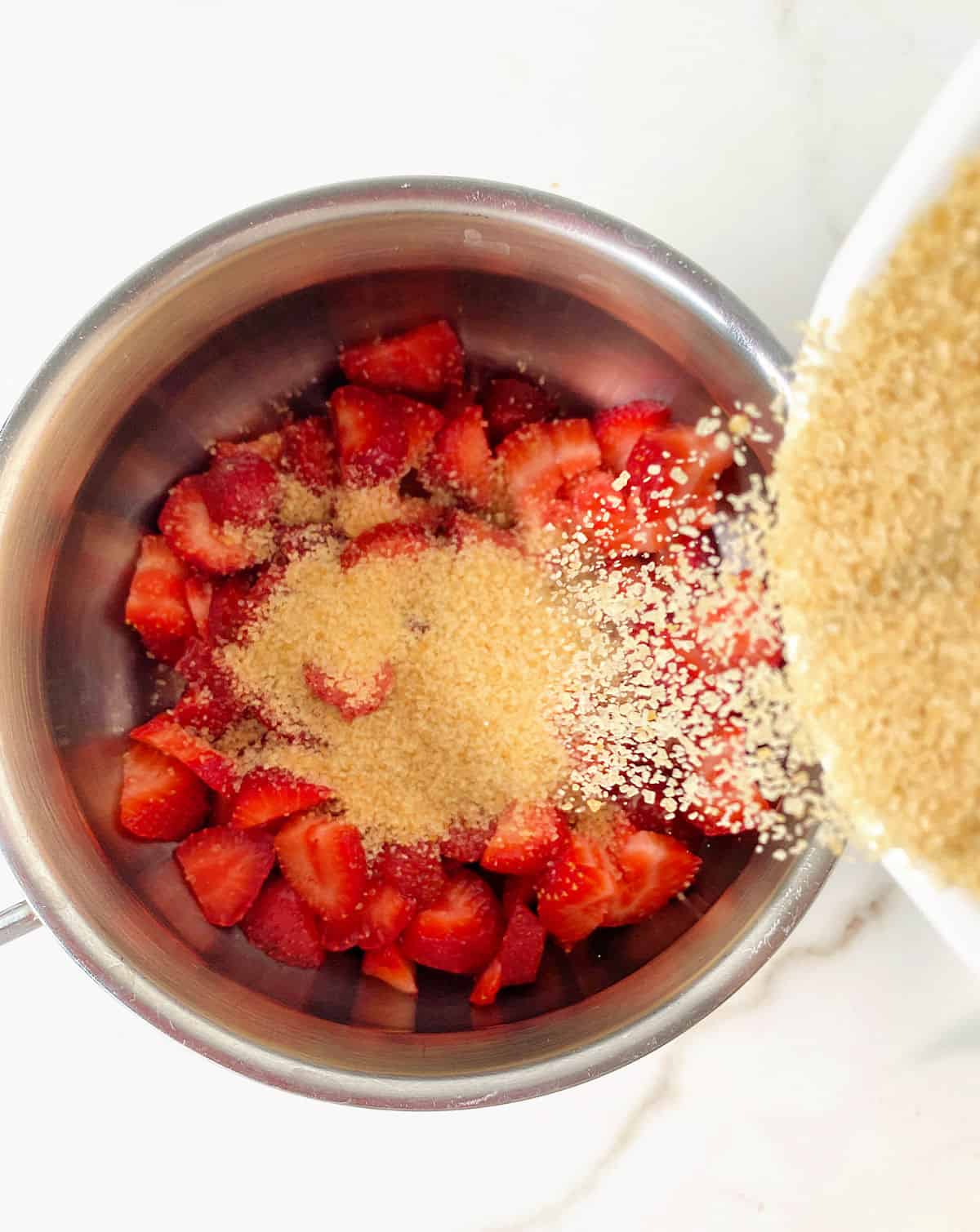 Adding light brown sugar to metal saucepan with chopped strawberries, white background