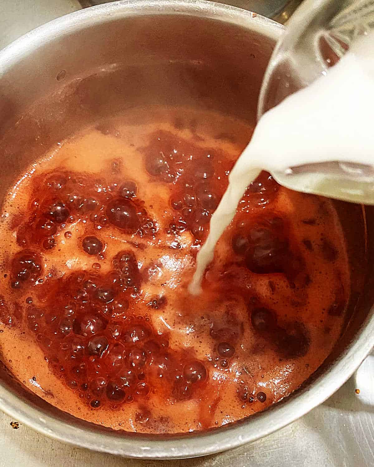Adding cornstarch slurry to a metal saucepan with boiling stawberry sauce.
